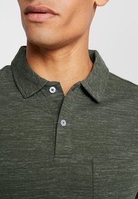 Tmavě zelený polo tričko vyrobené z texturovaného materiálu, s límečkem, dvěma knoflíky a kapsou na prsou. Přírodní vlákna s melírovaným vzhledem.