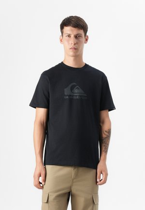 Un jeune homme aux cheveux courts se tient avec assurance dans un t-shirt noir arborant un logo, associé à un pantalon khaki, devant un fond blanc.