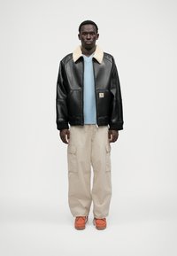 Czarna skórzana kurtka bomberka z kremowym sztucznym kołnierzem z barankiem, zestawiona z jasnoniebieską bluzką, beżowymi spodniami cargo i pomarańczowym obuwiem.