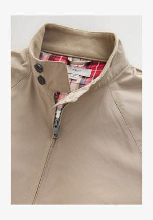 Beige Jacke aus leichtem Material mit Reißverschluss vorne, braunem, geripptem Kragen und rotem Kariervlies. Verfügt über zwei schwarze Knopfakzente.
