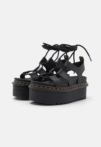 Svarte plattform sandaler laget av teksturert materiale, med krysset stropper og en tykk såle med gule sømmer langs kanten.