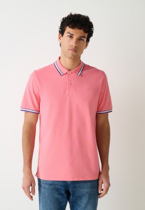 Giovane uomo con capelli ricci che indossa una polo rosa con colletto e polsini a righe blu e bianche e jeans blu, in piedi davanti a uno sfondo semplice.