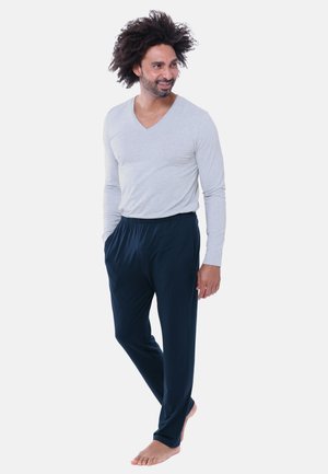 Man met krullend haar, die een lichtgrijs hemd met lange mouwen en donkerblauwe broek draagt, staat blootsvoets met zijn handen in de zakken.