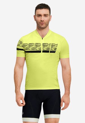 Mann steht und trägt ein gelbes kurzärmeliges Radtrikot mit schwarzen und weißen grafischen Streifen sowie schwarze Radhosen mit weißen Bändern.