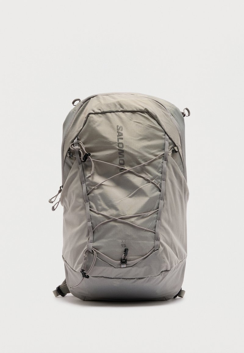 Ανοιχτό γκρι σακίδιο Salomon XT 15 με ελαστικά κορδόνια εμπρός, δίχτυα πλαϊνές θήκες και επάνω θήκη με φερμουάρ, όρθιο σε λευκό φόντο.