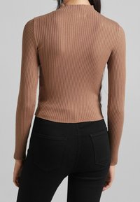 Femme portant un haut marron côtelé à manches longues ajusté, rentré dans un pantalon noir taille haute, vue de dos sur un fond gris uni.