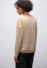 Beżowa bluza w luźnym fasonie, okrągłym dekolcie i ściągaczu na dole. Miękka tekstura, opadające ramiona i minimalistyczny design.