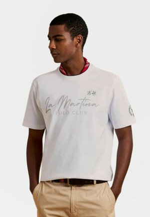 Jonge man die een wit La Martina Polo Club t-shirt draagt met beige broek, staat met handen in de zakken en kijkt naar opzij.