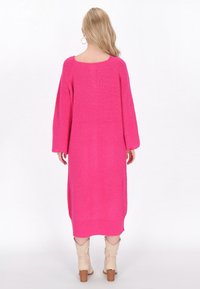 Svetlo roza pletena obleka z sproščenim krojem, širokimi rokavi in odprtim hrbtom. Teksturovana tkanina sega do sredine telesa in je kombinirana s bež čevlji.