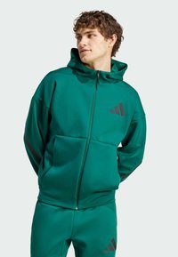 Zelena atletska kapuca z zadrgo na sprednji strani, dvema sprednjima žepoma in kapuco z vrvico. Ima črn Adidas logotip in črte na rokavih.