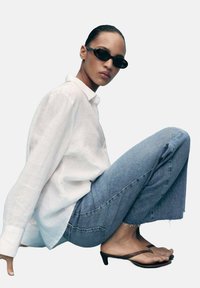 Bjela lanena košulja s dugim rukavima, svijetloplave poderane traperice, crne naočale za sunce na kopču i smeđe japanke s oštrim vrhom i niskom petom.