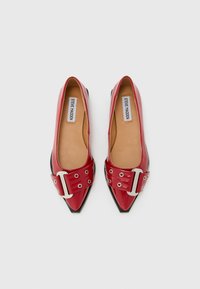 Une paire de ballerines en cuir verni rouge à bout pointu avec des détails de boucle en argent et des semelles beiges sur fond blanc.