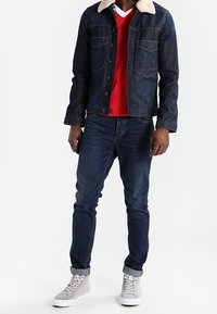 Denimjack met een beige schapenvachtkraag, een rode sweater eronder, blauwe jeans en grijze hoge sneakers met witte zolen.