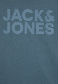 Modrá bavlnená tričko s veľkým svetlomodrým nápisom "JACK & JONES" na prednej strane. Hladká textúra s minimálnymi detailmi dizajnu.