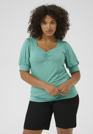 Top teal a maniche corte con scollatura quadrata, dettaglio arricciato al centro, abbinato a pantaloni corti neri. Tessuto morbido; consistenza liscia.