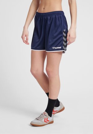 Osoba ubrana w granatowe sportowe szorty marki Hummel, czarne skarpetki i beżowe trampki z czerwonymi wzorami w kształcie litery V, stojąca na jasnoszarym tle.