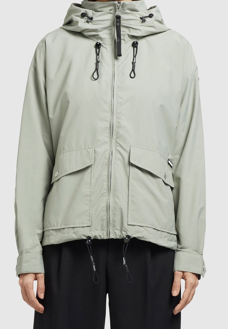 Veste à capuche gris clair avec fermeture éclair à l'avant, poches à rabat, cordons réglables à la capuche et à l'ourlet, portée sur un pantalon noir.