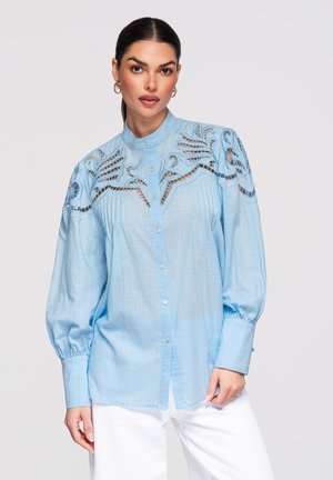 Kvinde iført en lyseblå bluse med knapper, broderede udskæringsdetaljer på skuldrene og puffede ærmer, kombineret med hvide bukser.