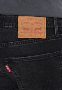 Dos d'un jean Levi's noir montrant un patch de marque en cuir et une étiquette rouge Levi's sur la poche, porté avec une chemise grise.