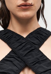 Gros plan du bas du visage et du cou d'une femme portant un vêtement en tissu noir froncé avec de larges bretelles croisées.