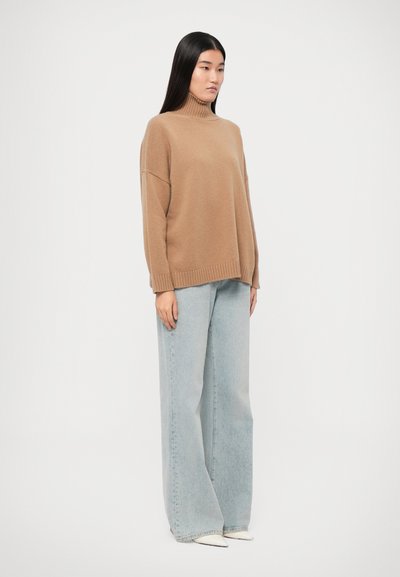 Pull beige surdimensionné avec un col haut, associé à un jean large bleu clair. Texture lisse et coupe décontractée dans un style casual.