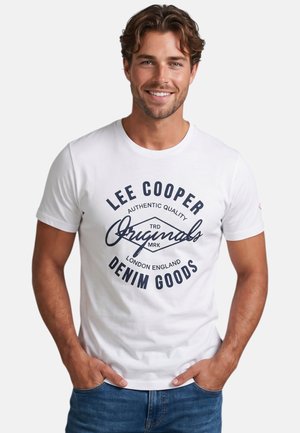 Homme souriant aux cheveux bruns portant un t-shirt graphique blanc Lee Cooper et un jean bleu, debout les mains dans les poches.