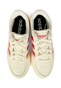 Baskets créme avec des rayures en cuir rouge, bout rond, lacets à l'avant et semelle en caoutchouc texturé. Logo "adidas" sur la languette et la semelle intérieure.