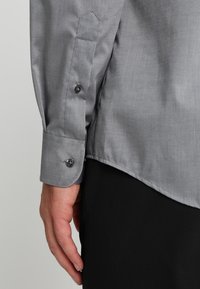 Szara koszula z długim rękawem o gładkiej fakturze, z mankietami zapinanymi na guziki i lekko zaokrąglonym dołem, zestawiona z czarnym materiałem pod spodem.