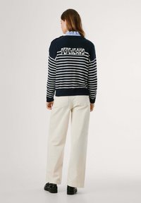 Maglione a righe navy e bianche con texture a maglia, caratterizzato da un logo sulla schiena, abbinato a pantaloni a gamba larga crema e scarpe nere.