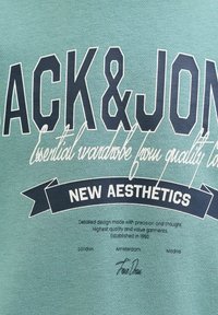 Vihreä kangas, jossa on mustavalkoinen teksti "JACK & JONES", kursiivinen lause ja lippu, jossa lukee "NEW AESTHETICS" pienemmillä paikkakuntien nimillä alla.
