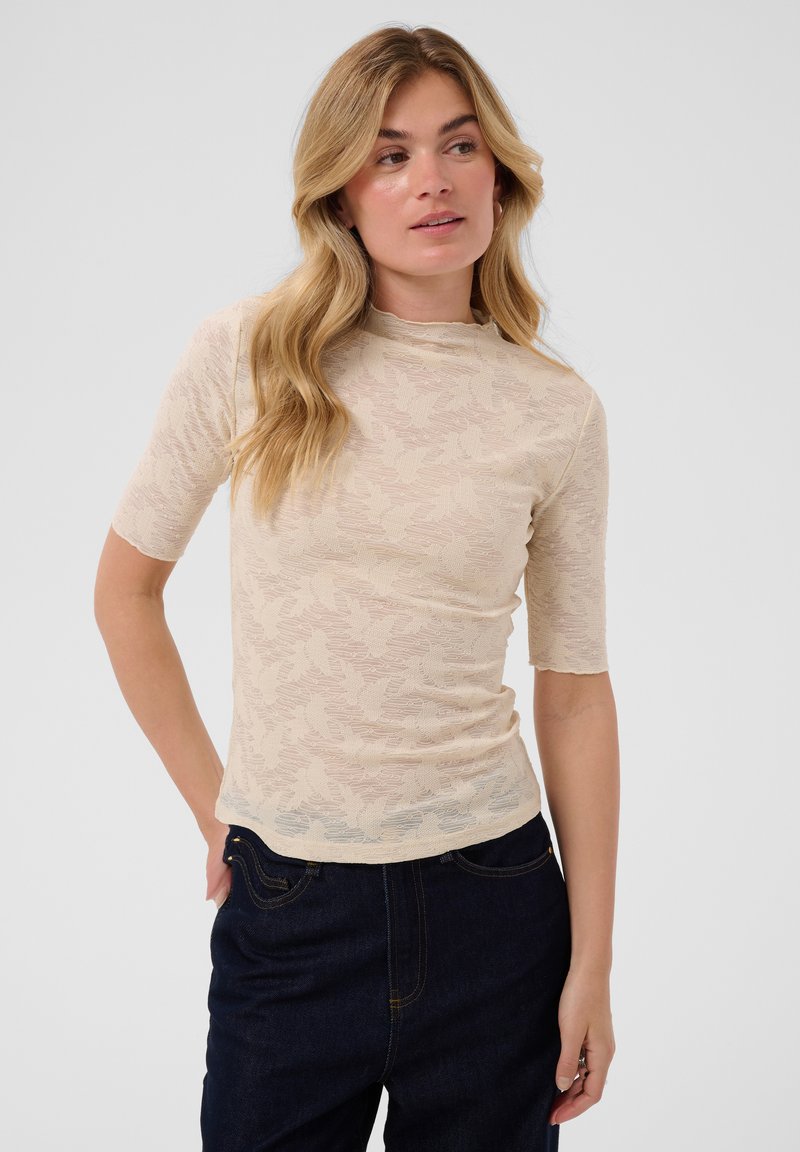 Femme aux longs cheveux blonds porte un haut ajusté à manches courtes, beige clair à motifs, et un jean foncé, debout avec une main dans sa poche.
