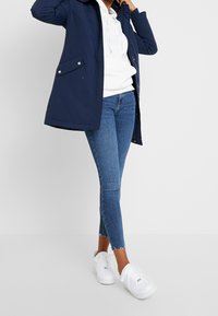 Navy blue waterproof jacket with a hood, slim-fit white hoodie underneath, distressed blue jeans, and white sneakers.