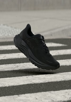 Schwarzer Hoka-Laufschuh schwebt in der Luft über einem nassen Zebrastreifen, wobei Wassertropfen von der Sohle spritzen.