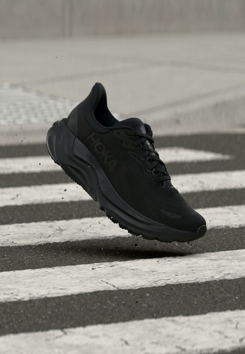 Μαύρο παπούτσι τρεξίματος Hoka στον αέρα πάνω από μια βρεγμένη διάβαση πεζών με σταγόνες νερού που πετάγονται από τη σόλα.