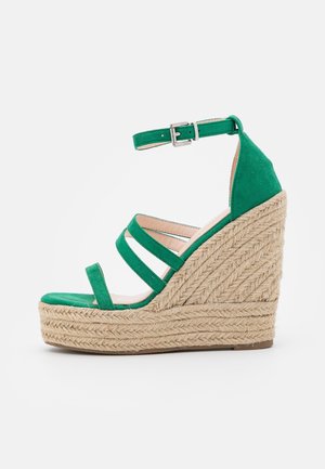 Sandale compensée en daim vert avec bride cheville et boucle argentée, semelle plateforme en jute tressé, et trois brides sur le devant au-dessus de l'orteil ouvert.
