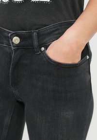 Main dans la poche avant d'un jean en denim noir avec un bouton visible et des passants de ceinture, portant un haut foncé.