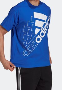 Modrá bavlněná tričko s kulatým výstřihem. Na přední straně je velké bílé logo Adidas a geometrické vzory. Krátké rukávy, pohodlný střih.