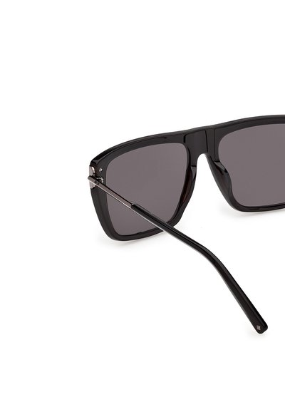 Gafas de sol rectangulares negras con lentes oscuros y varillas delgadas y curvas que se extienden hacia la derecha sobre un fondo blanco.