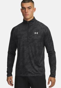 Zwart atletisch shirt met lange mouwen en een halve rits kraag. Beschikt over een camouflagepatroon, lichtgewicht stof en een klein wit logo op de borst.