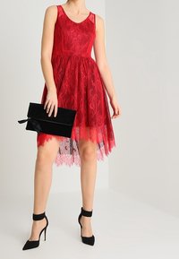 Femme portant une robe rouge en dentelle sans manches tenant une pochette noire, associée à des escarpins noirs à bride à la cheville, debout devant un fond uni.