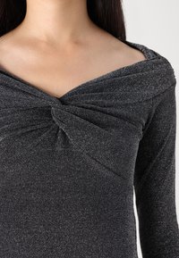 Schwarzes Langarmoberteil mit einem verdrehten Knoten-Design am Ausschnitt und einer schimmernden Textur, die eine figurschmeichelnde Silhouette bietet.