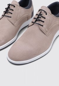 Zapatos de cuero texturizado en color beige con acentos negros, suela blanca y cordones negros. El diseño presenta perforaciones y una silueta elegante.