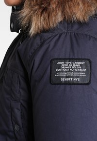 Marinfärgad parka med en huva kantad av päls. Har en svart patch med vit text. Materialet verkar slätt, med knappar på sidan.