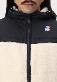 Mustassa ja kermahuivissa on pörröinen, teksturoidusta alaosasta, sileä yläosa ja edessä vetoketju säädettävällä hupulla. K-Way-logo näkyvissä.