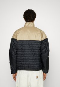 Chaqueta acolchada con una sección superior beige y la mitad inferior negra. Cuenta con un cuello alto y textura acanalada en la zona negra.