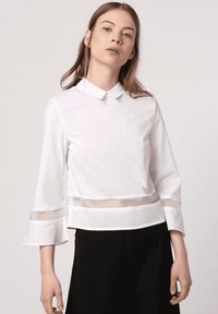 Blusa blanca de cuello con paneles transparentes, mangas acampanadas de tres cuartos y un diseño cropped. Textura suave, con un detalle mínimo.