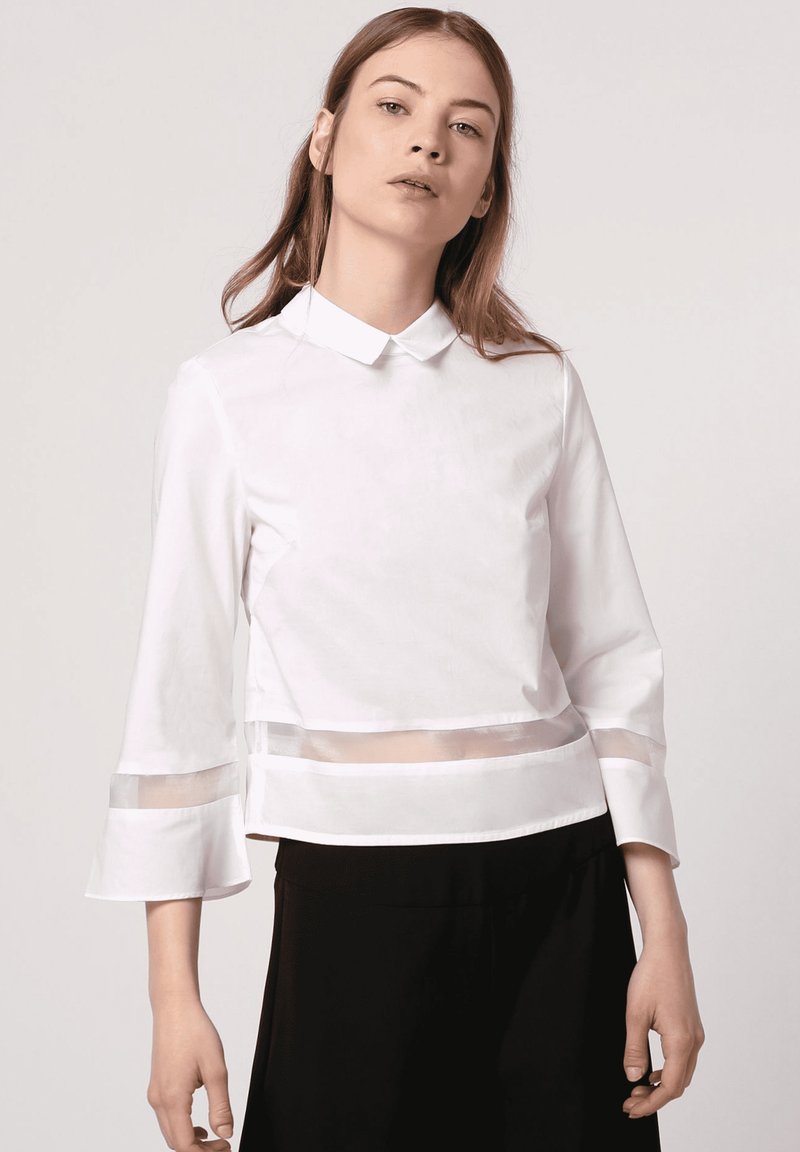 Blusa blanca de cuello con paneles transparentes, mangas acampanadas de tres cuartos y un diseño cropped. Textura suave, con un detalle mínimo.