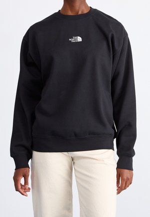 Osoba ubrana w czarną bluzę North Face z długim rękawem i jasnobeżowe spodnie na jednolitym jasnym tle.
