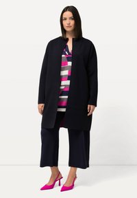 Lång svart kappa med rak skärning, bärs över en randig topp i rosa, grå och marinblå, kombinerad med marinblå culottes och klara rosa klackar.