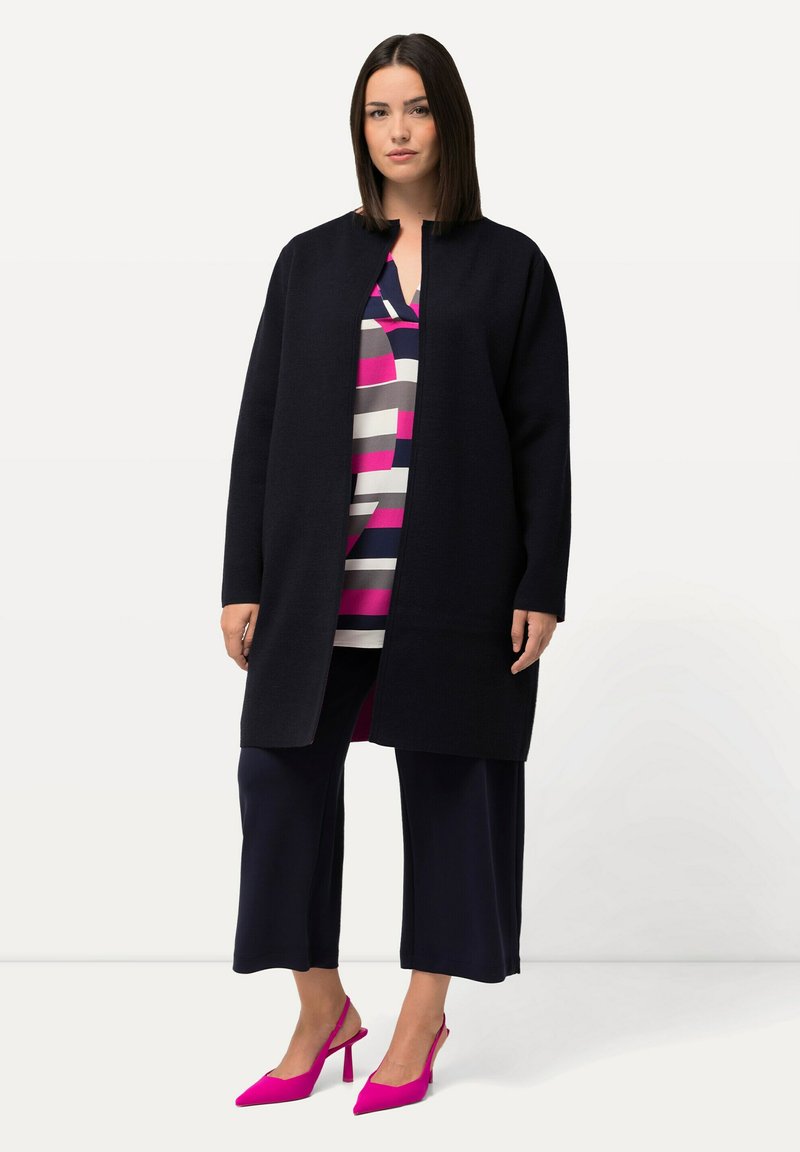 Lång svart kappa med rak skärning, bärs över en randig topp i rosa, grå och marinblå, kombinerad med marinblå culottes och klara rosa klackar.